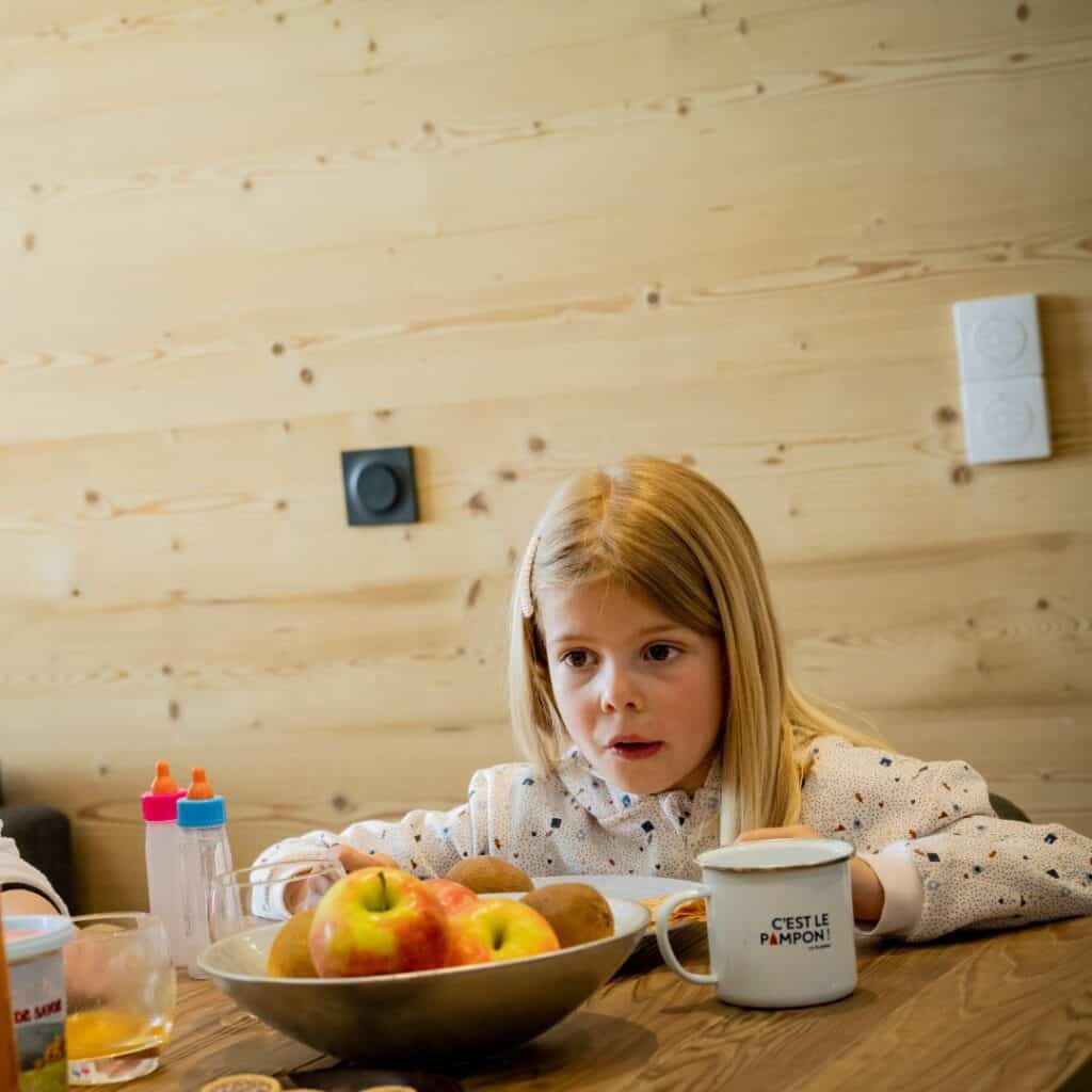 A young girl sits at a wooden dining table with fruit and mugs, starting the day with a relaxed breakfast in a self catering La Plagne apartment.