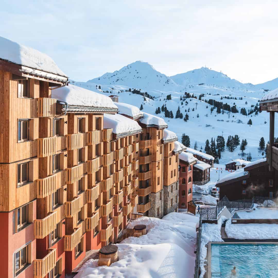 Wooden self catering La Plagne chalets with balconies overlook a scenic mountain backdrop, with a heated outdoor pool visible below.