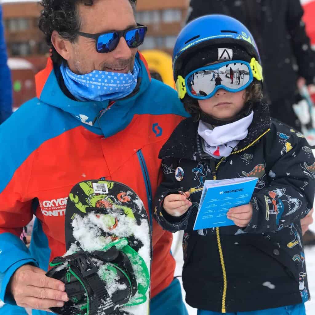 A young child in a colorful snow jacket and blue helmet stands holding a certificate from Oxygène, a ski and snowboard school, next to an Oxygene instructor in bright red and blue gear holding a snowboard covered in snow. This moment captures the end of a snowboarding lesson for kids in La Plagne ski resort.