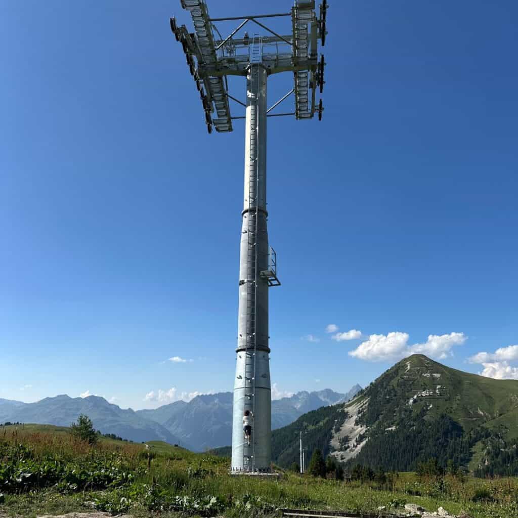 A tall gondola lift tower stands against a clear blue sky, with a worker climbing the base and alpine mountains in the distance. The lush green slope and rugged peaks suggest a high-altitude location under development.