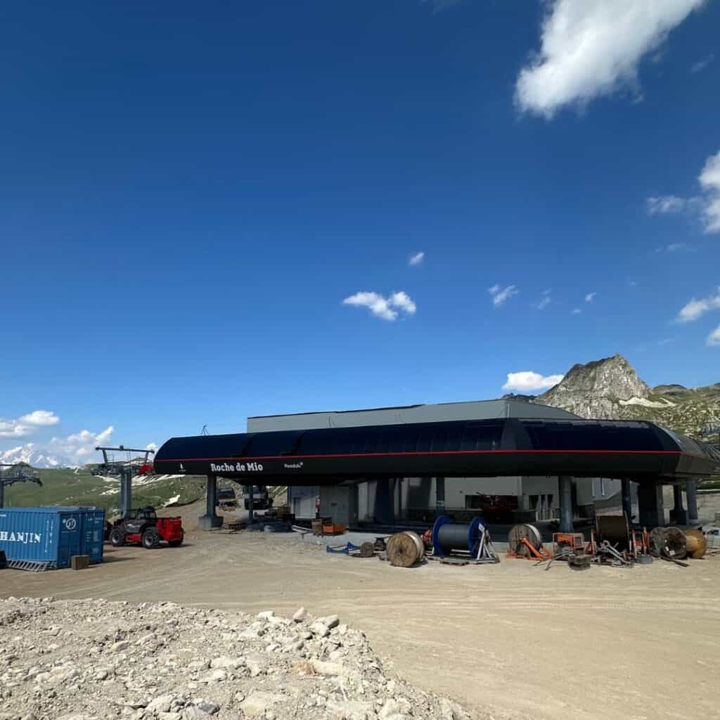 A wide view of the gondola mid-station base at Cole de Forcle, labeled “Roche de Mio,” surrounded by construction equipment and cable spools. The structure sits on dusty terrain with scenic mountain ridges in the background.