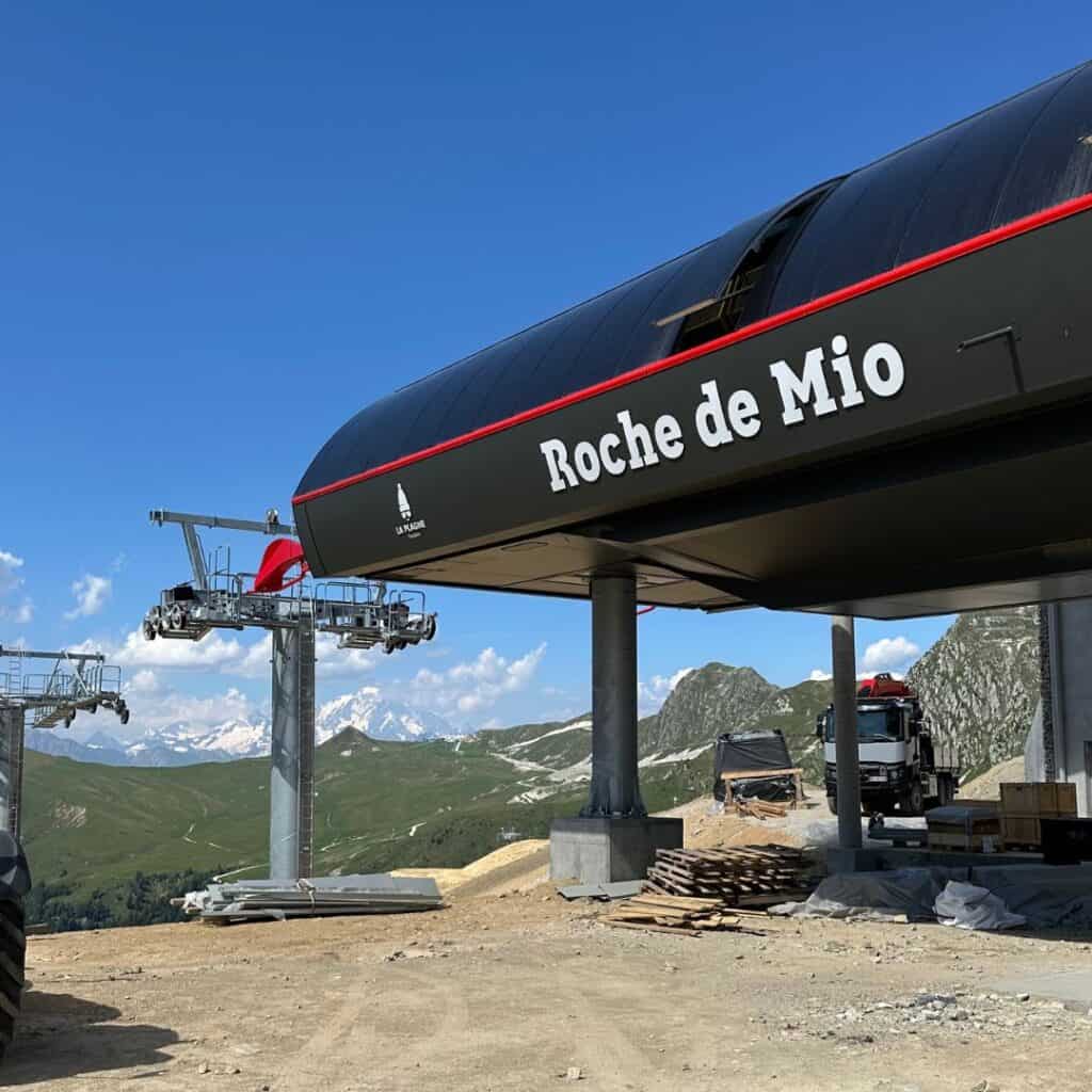 Close-up of the “Roche de Mio” gondola station with sleek modern architecture, ski lift equipment, and views of snow-capped peaks in the distance. Construction materials are scattered nearby, signaling active installation.