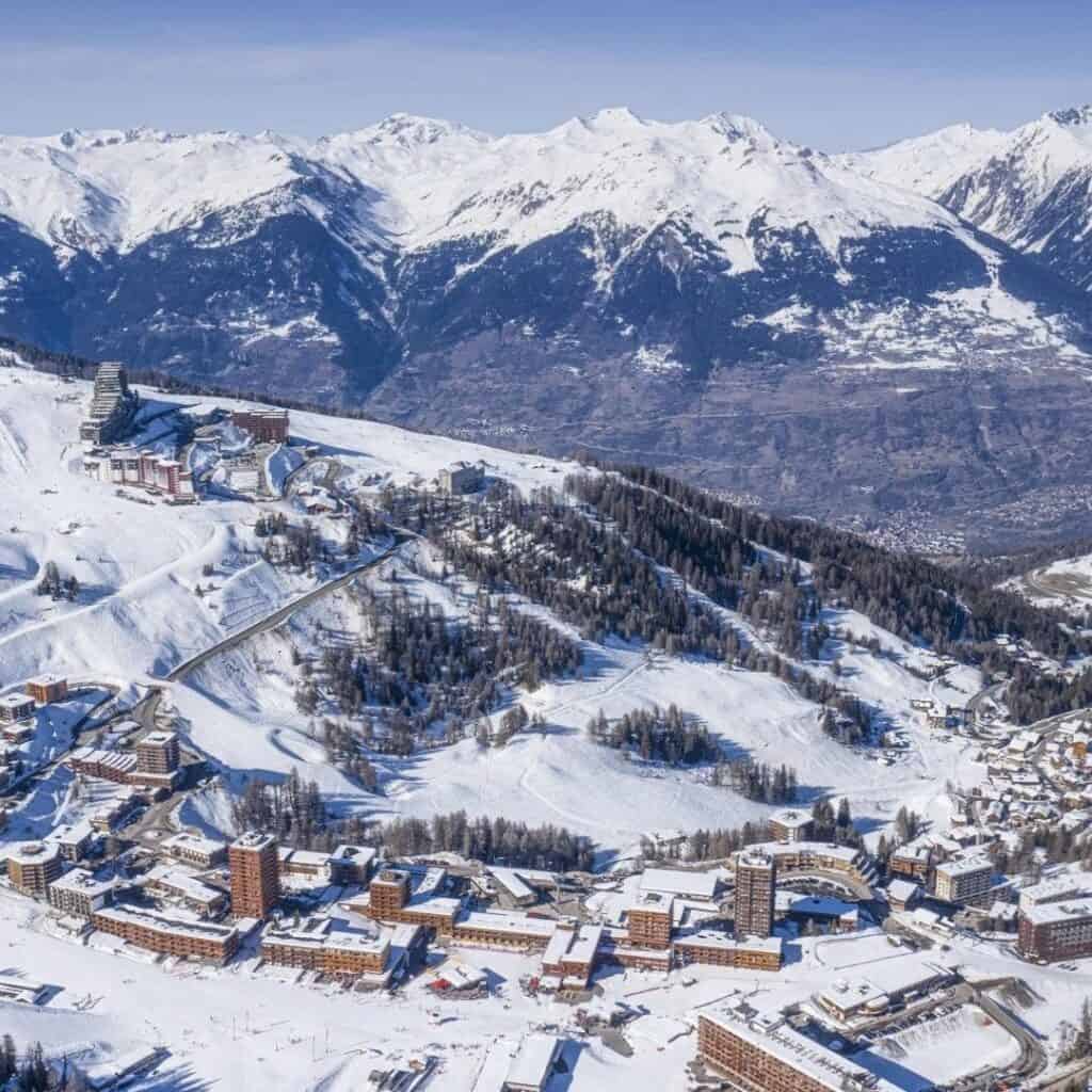 Wide aerial shot of La Plagne covered in snow, showing clusters of ski resort buildings nestled among winding ski runs and framed by towering snow-covered Alps in the background.