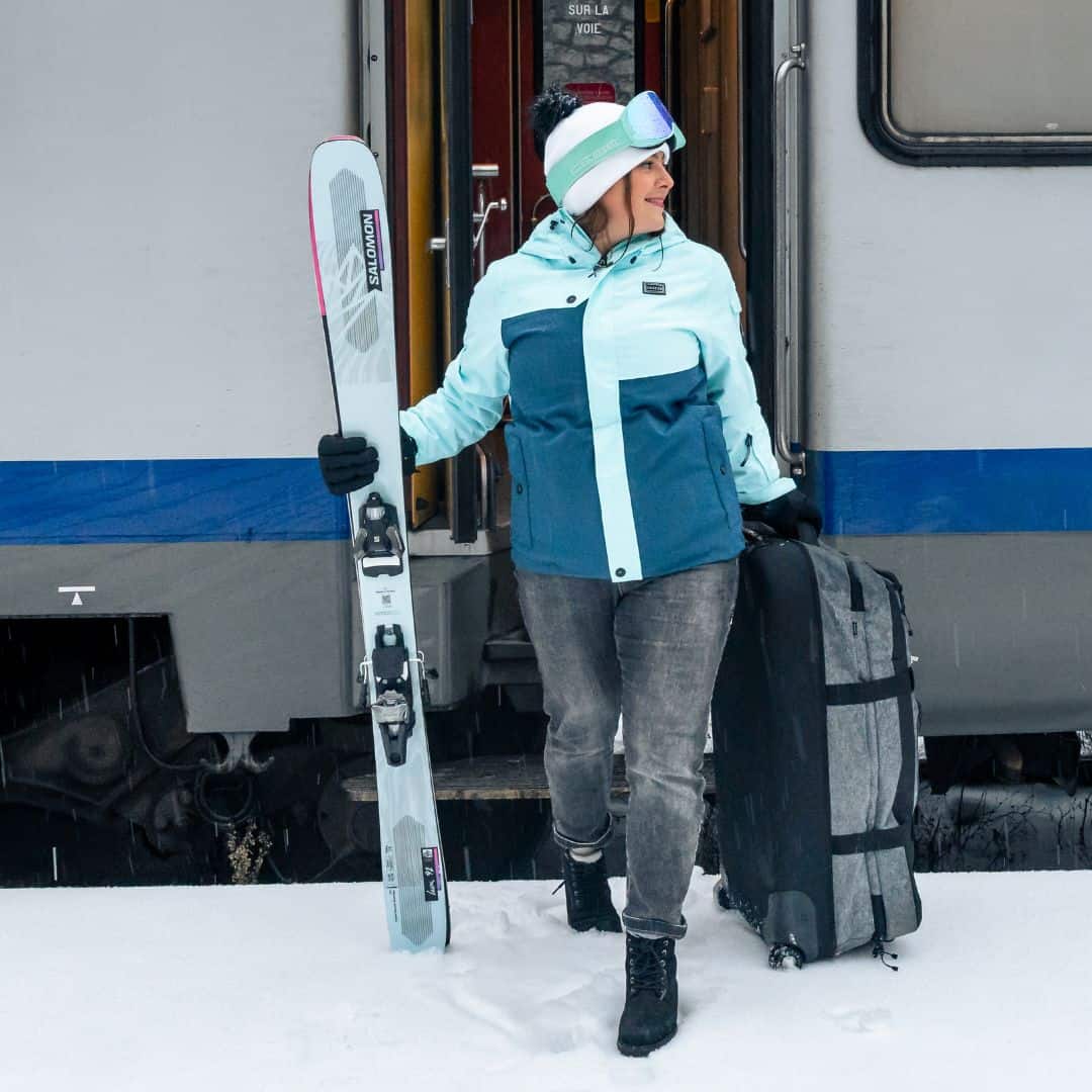 A woman steps off a train onto snowy ground, holding skis in one hand and pulling a large suitcase, ready to ski at La Plagne.