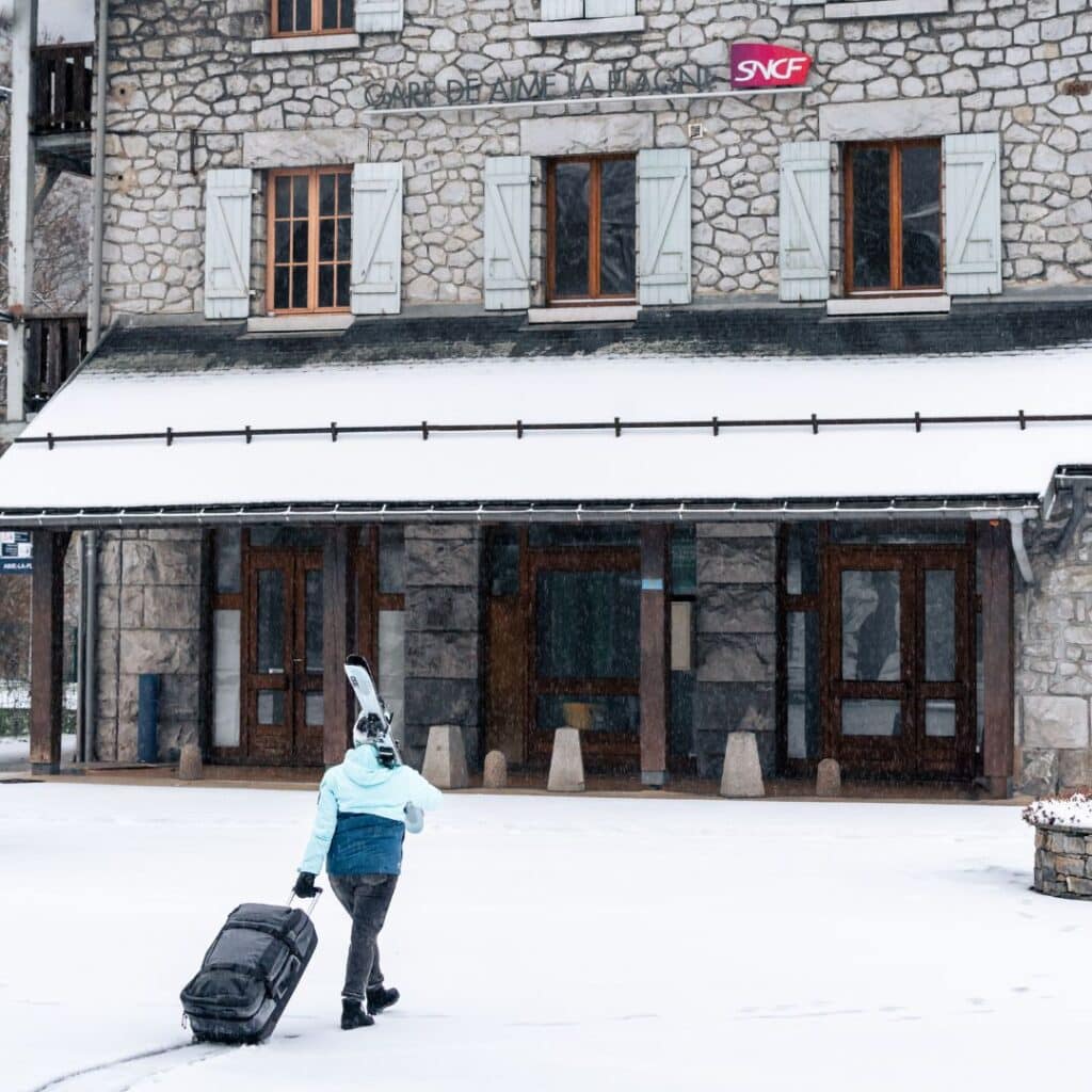 A person in a light blue ski jacket walks through fresh snow outside Gare d’Aime La Plagne SNCF station, carrying skis and luggage.