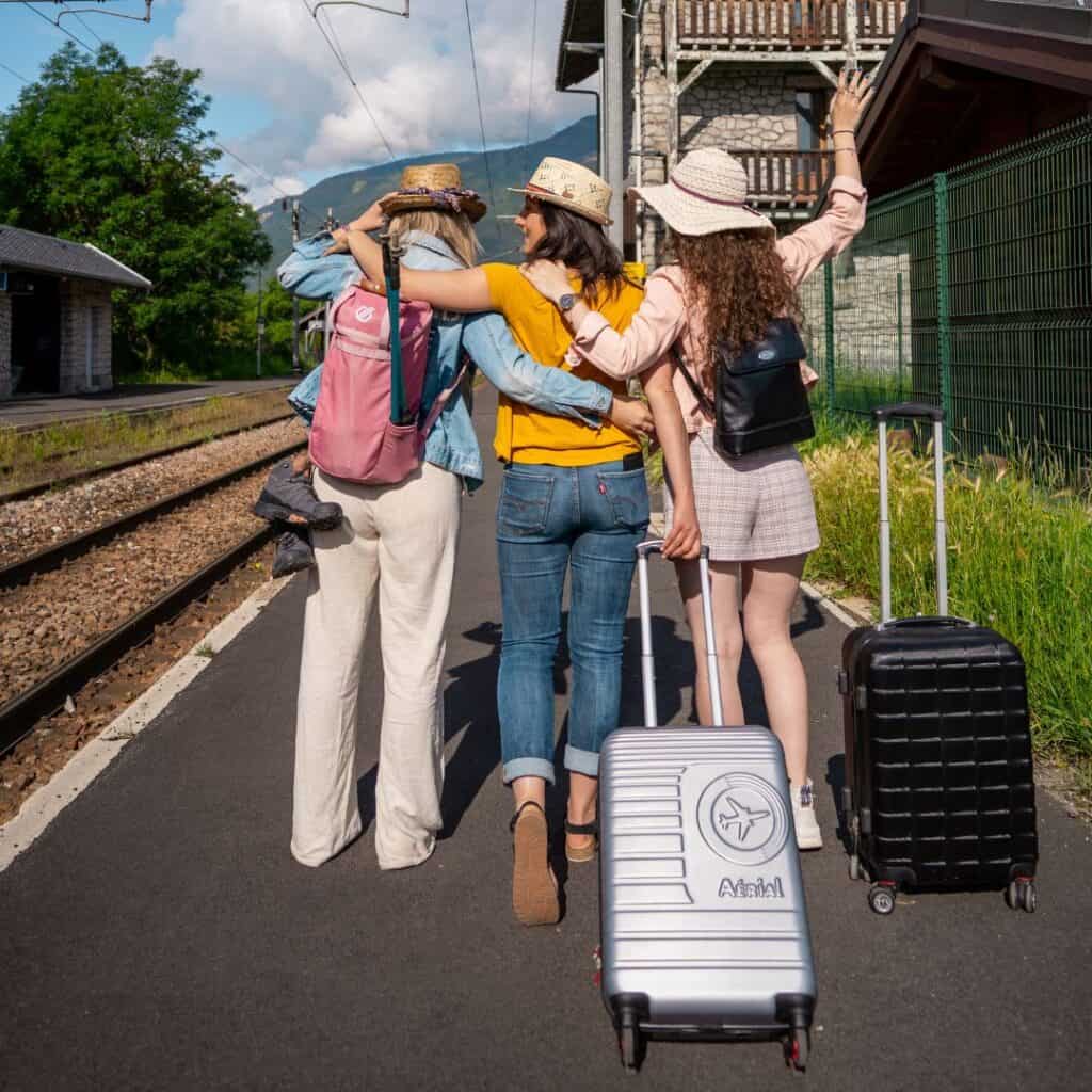 Three friends with hats and suitcases walk arm-in-arm down a sunny train platform toward the tracks, excited for a mountain getaway.