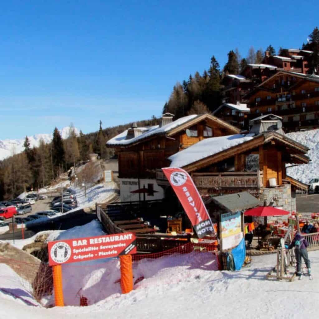 Rustic bar restaurant with wooden exterior and signage reading “Bar Restaurant” offering Savoyard specialties, with ski gear and visitors nearby under a clear blue sky.