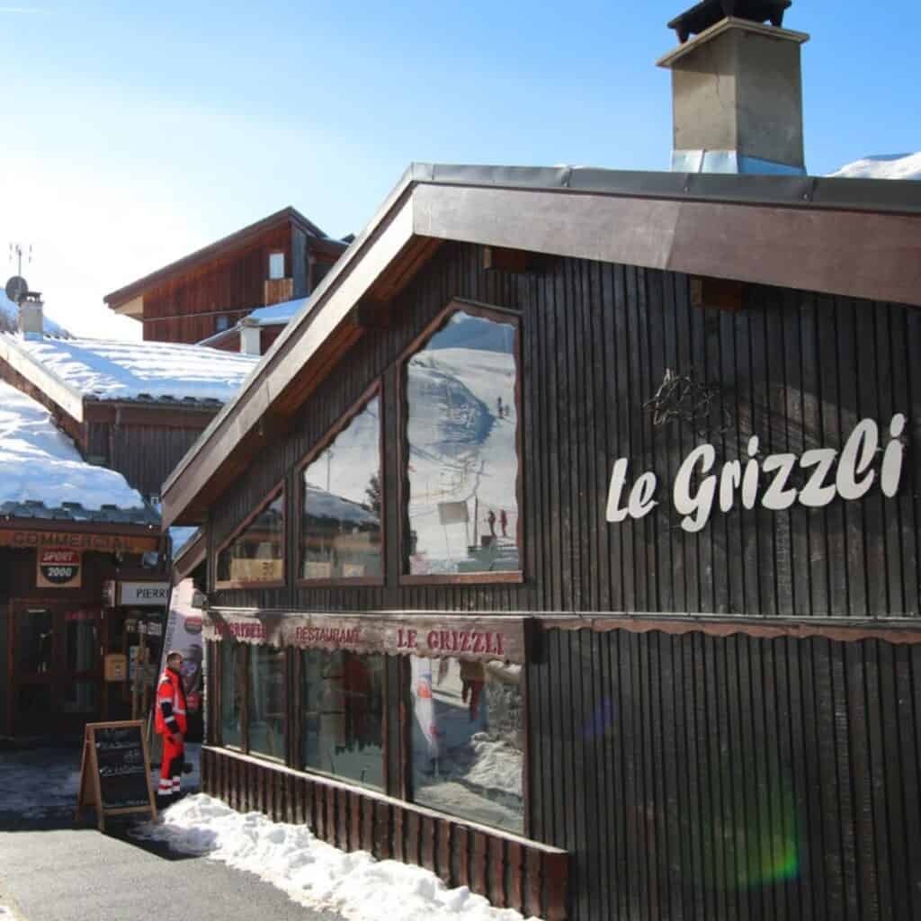 Street view of Le Grizzli restaurant with large glass windows reflecting nearby ski slopes, wooden paneling, and a visible sign above the entrance.