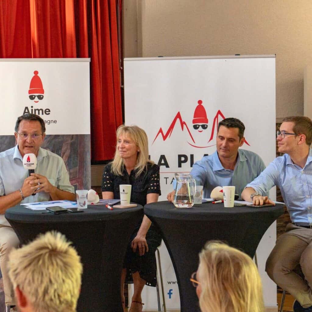 Four panelists speak during a town hall meeting for the Aime La Plagne gondola proposal, with La Plagne banners in the background highlighting the new lift La Plagne development.