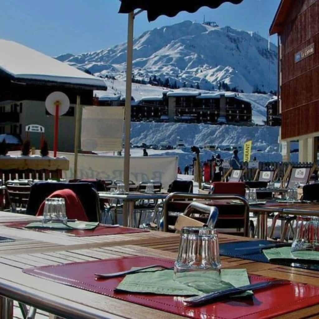Tables set with placemats and glasses at an outdoor terrace dining area, with a snowy mountain peak and ski resort buildings visible in the background.