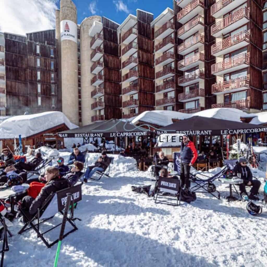 Crowded snow-covered patio of Le Capricorne bar and restaurant, with skiers lounging in sun chairs at the base of a tall wooden hotel building marked “La Plagne.”