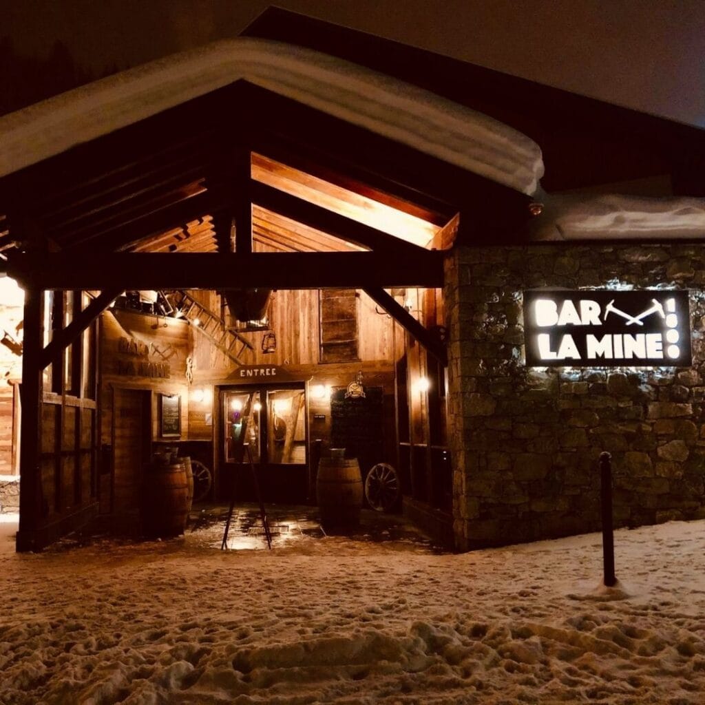 Warmly lit entrance to Bar La Mine at night, featuring a stone and wood exterior, rustic décor, and a glowing sign with a crossed pickaxe design.