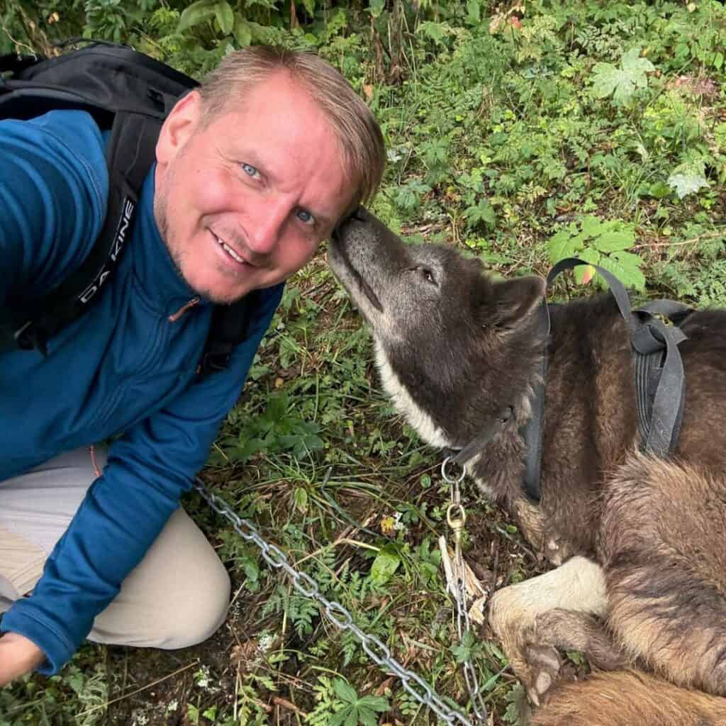 A man kneels next to a laying sled dog who affectionately sniffs his ear. Captured during a cani rando La Plagne adventure, the bond between human and husky shines through.