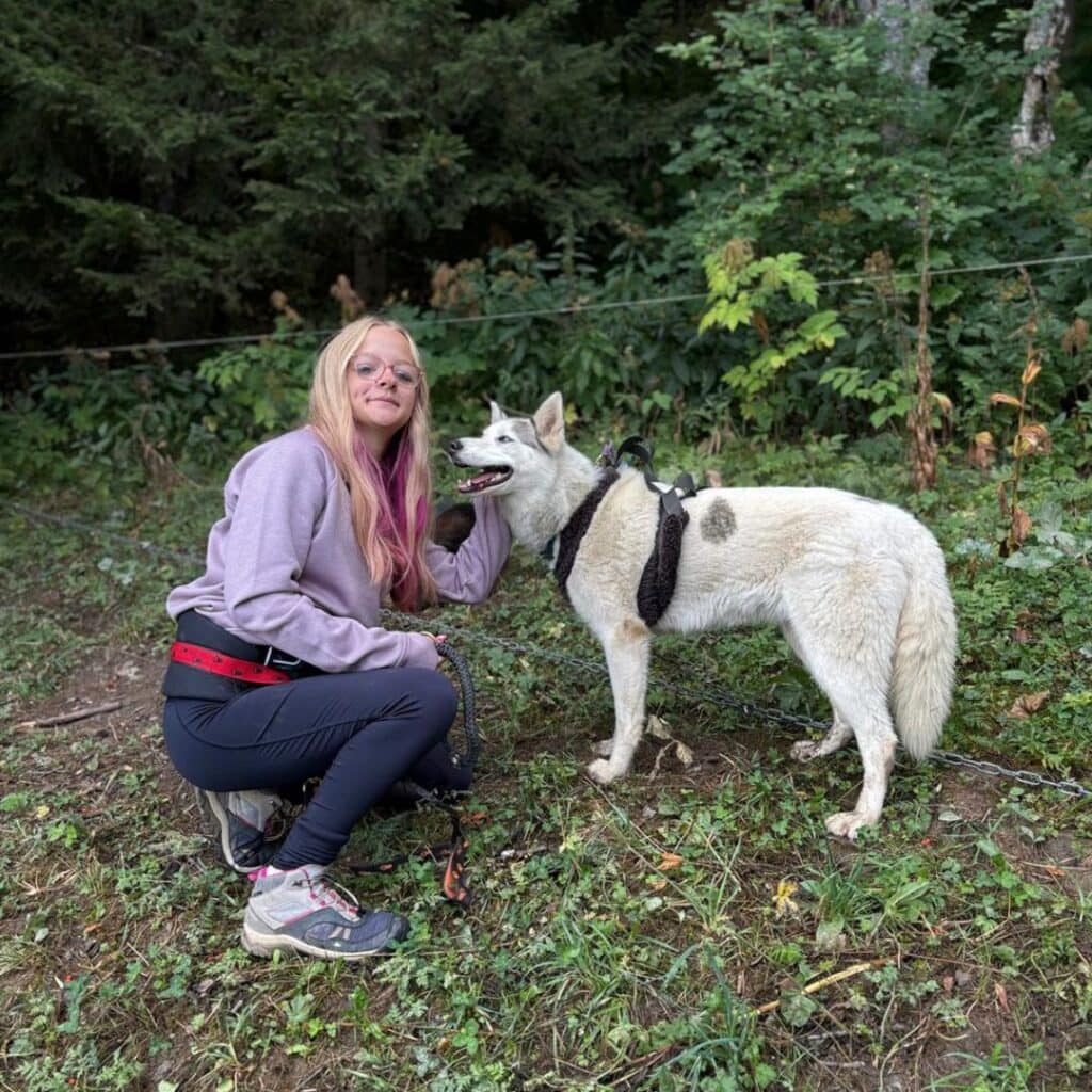 Teen girl crouching beside a white husky with black spots, both looking cheerful. Taken during a family-friendly cani rando La Plagne activity in the forest.