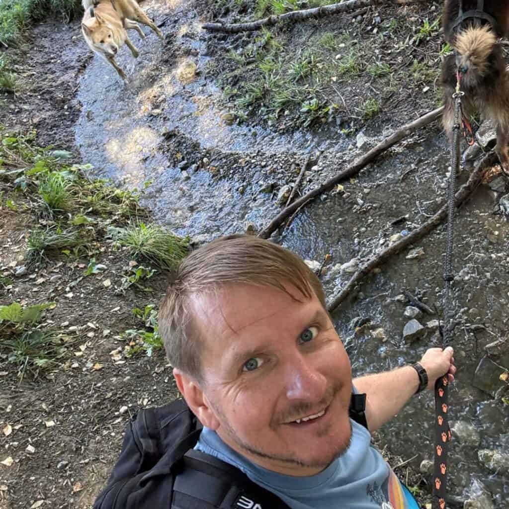 Man smiling for a selfie while leading a husky across a narrow woodland stream, another dog bounding in the background. Playful moment from an adventurous cani rando La Plagne hike.