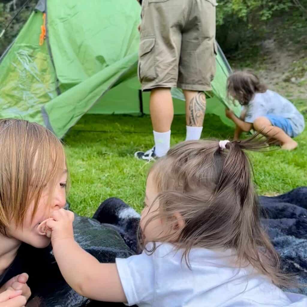 A toddler reaches out to feed a snack to another child lying on a blanket, while two other family members set up a tent in the background.