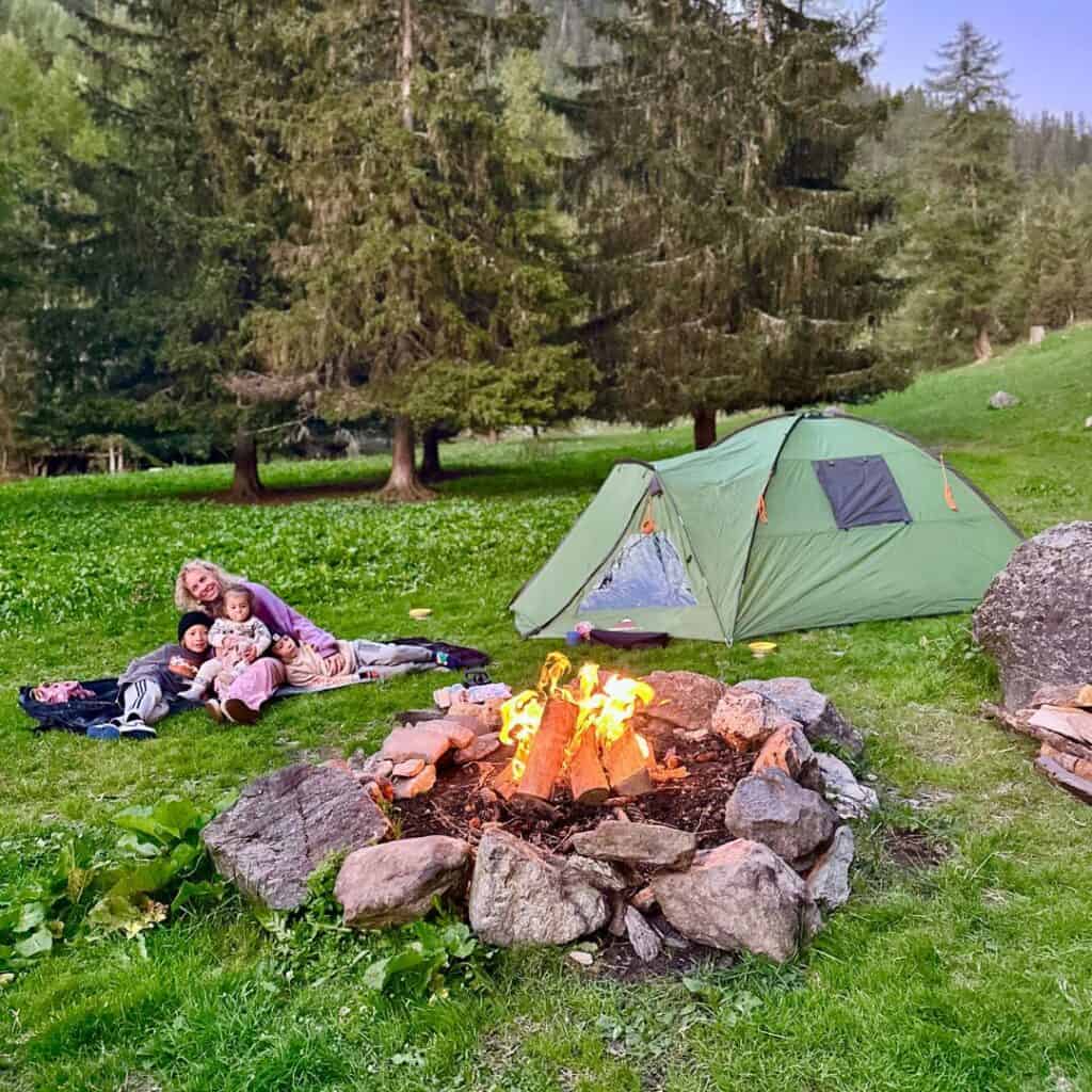 A woman sits on a blanket with two children beside a campfire ring, with a green tent pitched nearby and tall pine trees in the French Alps.