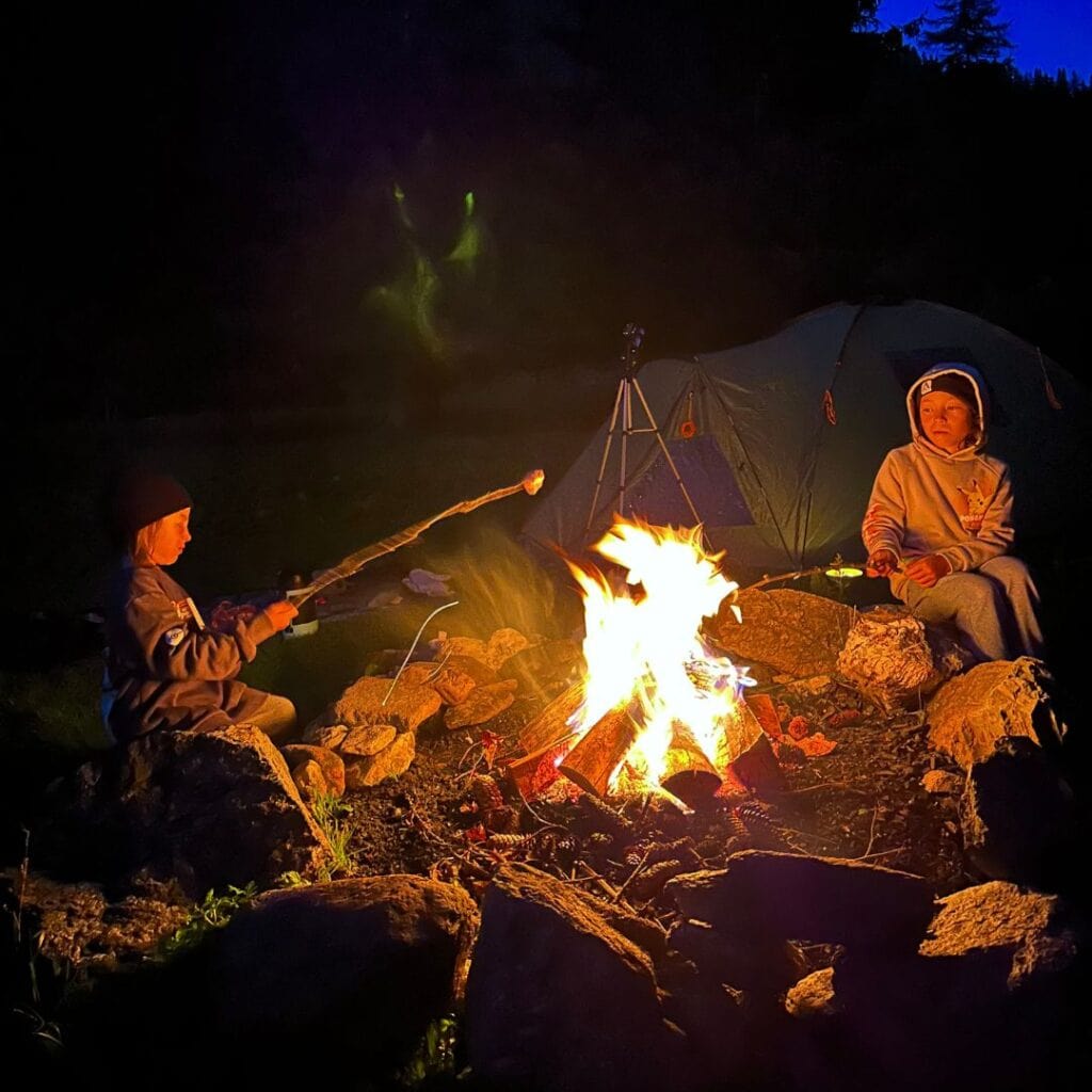 Two children sit by a glowing campfire at night, roasting marshmallows on long sticks, with a tent set up in the background during camping in La Plagne, French Alps.