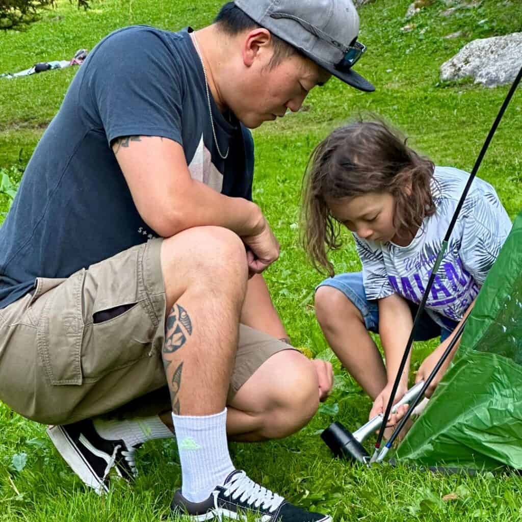 An adult and a child work together to hammer tent stakes into the ground while setting up camp on green grass in La Plagne, French Alps.
