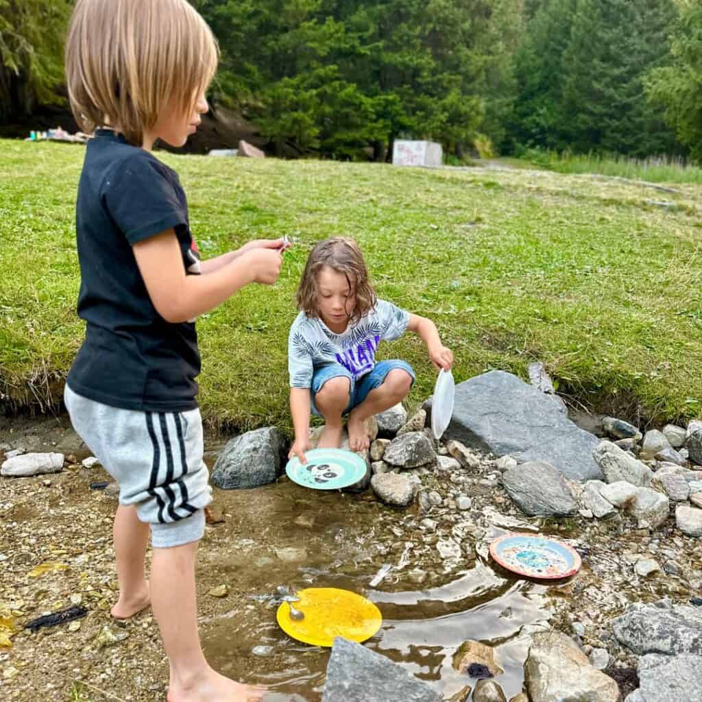 Two barefoot children wash colorful camping plates in a shallow rocky stream surrounded by alpine meadow.