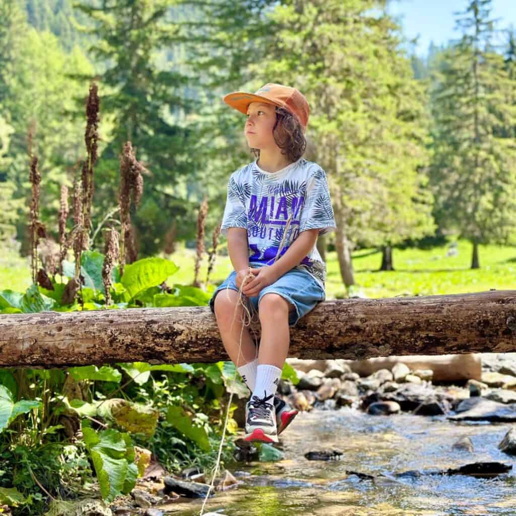 A child wearing an orange cap sits on a fallen log over a stream, holding a fishing line, with alpine forest scenery in La Plagne behind.