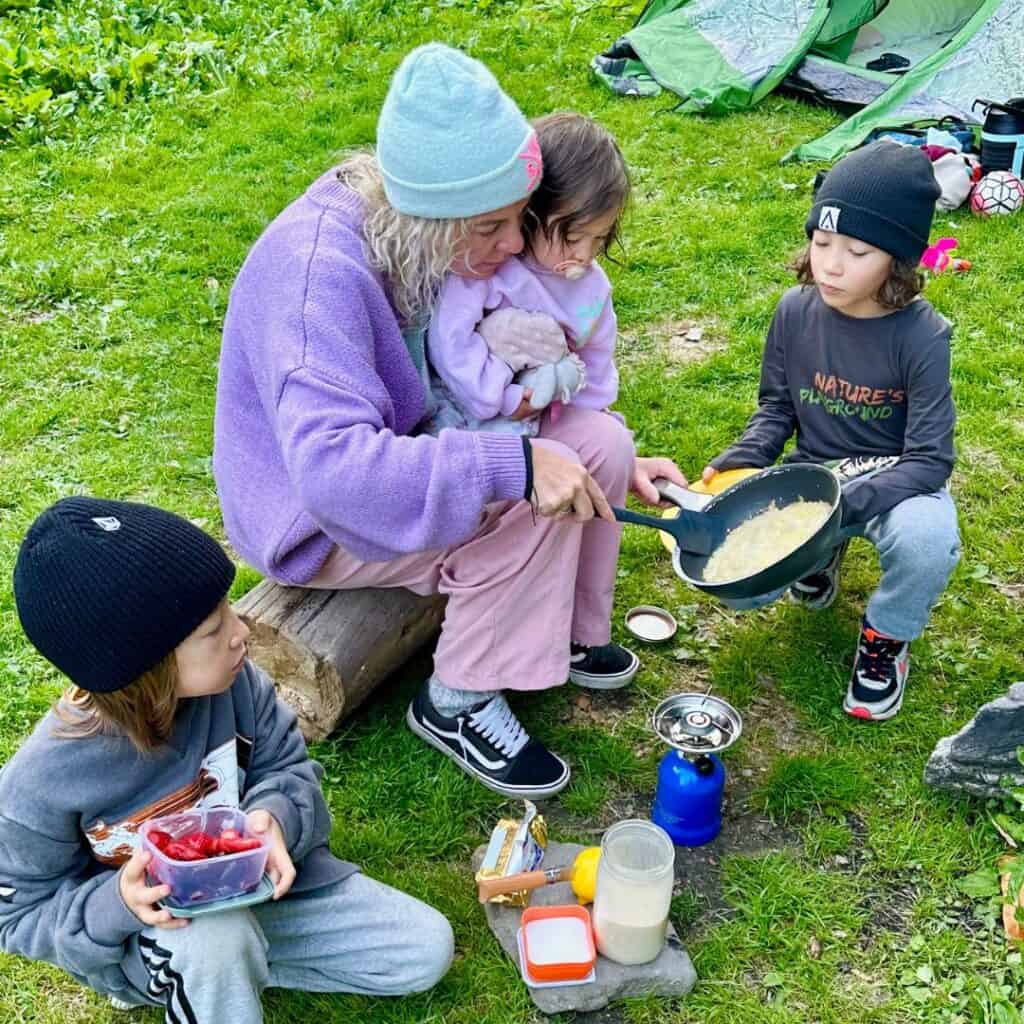 A woman in a purple sweater cooks in a frying pan on a small camping stove while two children watch and one sits on her lap, with tents in the background.