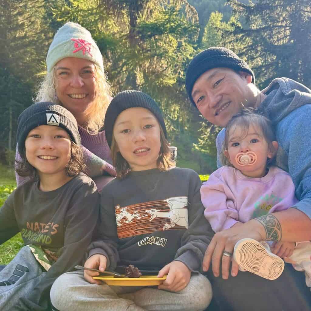 A family of five wearing warm layers and beanies smiles at the camera while sitting together outdoors during a sunny morning in La Plagne.