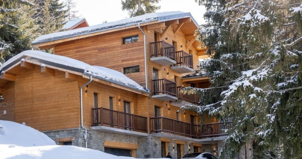 Snow-covered exterior of Chalet du Bois Melay, a luxury chalet in La Plagne, with wooden balconies and stone foundations nestled among tall pine trees.