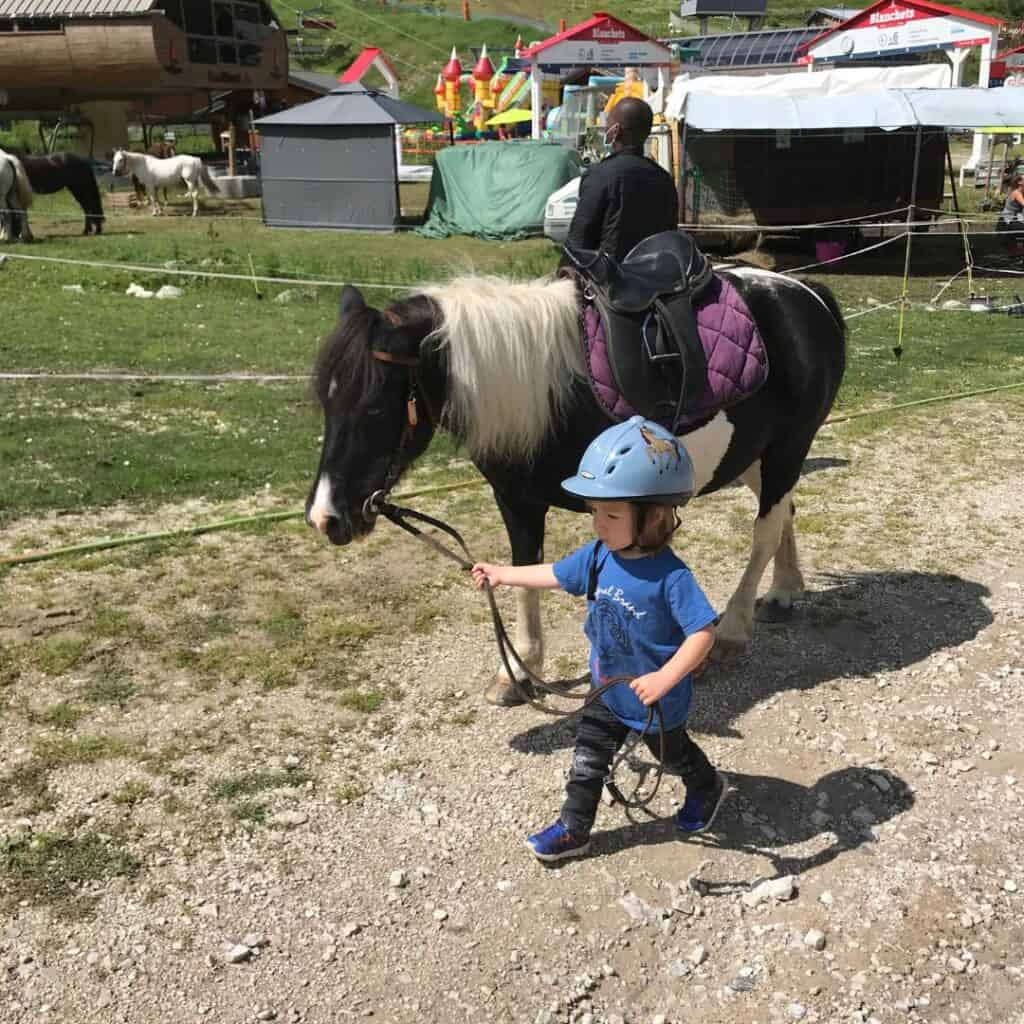 A young child wearing a helmet confidently leads a black and white pony with a purple saddle pad at a sunny equestrian area in La Plagne. In the background are other horses and colorful structures, indicating a fun family-friendly setup.