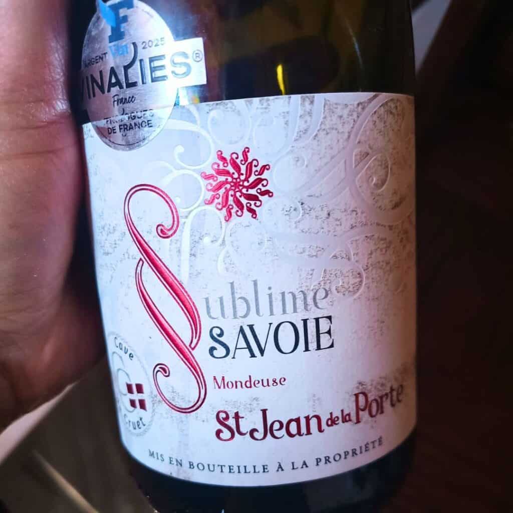Close-up of a bottle of “Sublime Savoie Mondeuse” wine from St Jean de la Porte. The label is ornate with red and silver details, showcasing a regional wine enjoyed at Chalet des Vignobles.