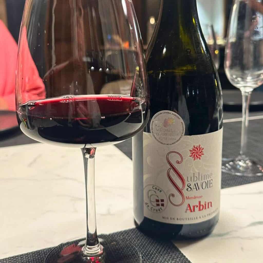 A glass of deep red Mondeuse wine sits beside a bottle of Sublime Savoie Arbin from Cave de Cruet. The elegant setup on a marble table reflects the refined local wine experience at Chalet des Vignobles.