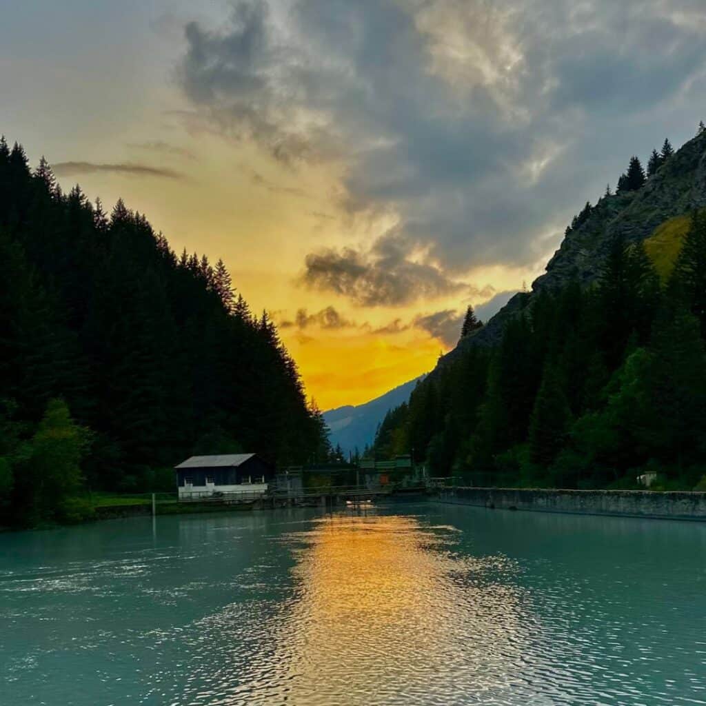 A golden sunset reflects off a calm river surrounded by dense pine forest and mountains near La Plagne. A small wooden cabin sits at the water's edge, adding to the tranquil alpine setting.