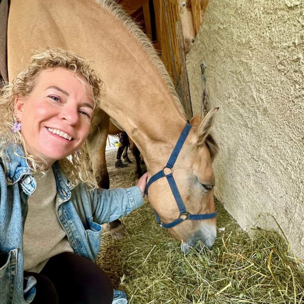 A smiling woman crouches next to a light brown horse as it eats hay inside a rustic stable near Chalet des Vignobles. The close bond between rider and horse is evident.