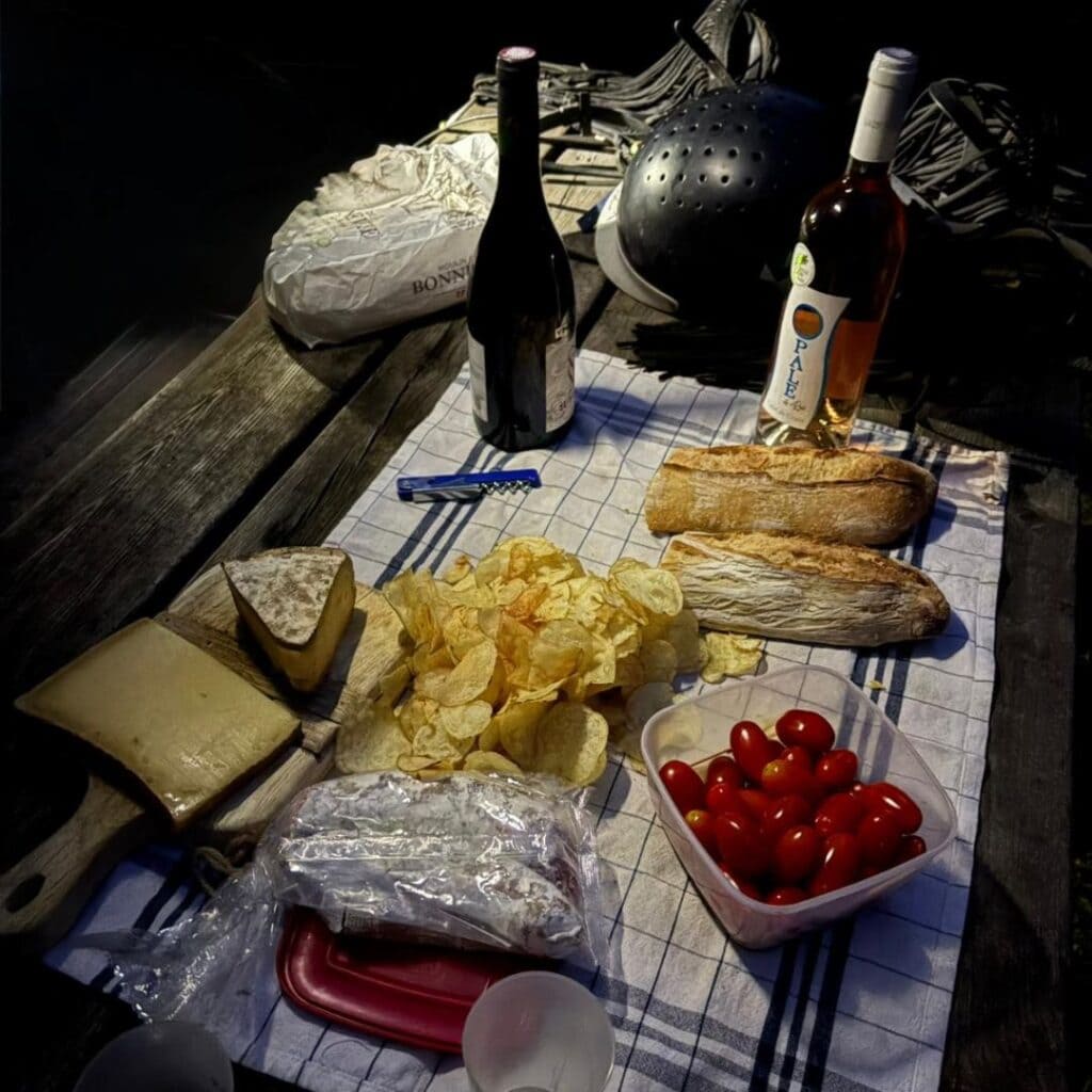 A rustic picnic on a checkered cloth featuring French bread, cheese, cherry tomatoes, potato chips, and two bottles of wine. Horse-riding helmets and gear in the background hint at a post-ride meal near Chalet des Vignobles.