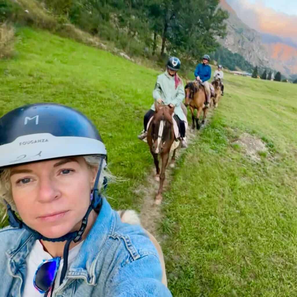 A woman in a riding helmet takes a selfie while leading a line of horseback riders through a green alpine meadow. Behind her, riders follow single file with mountain peaks in the distance.