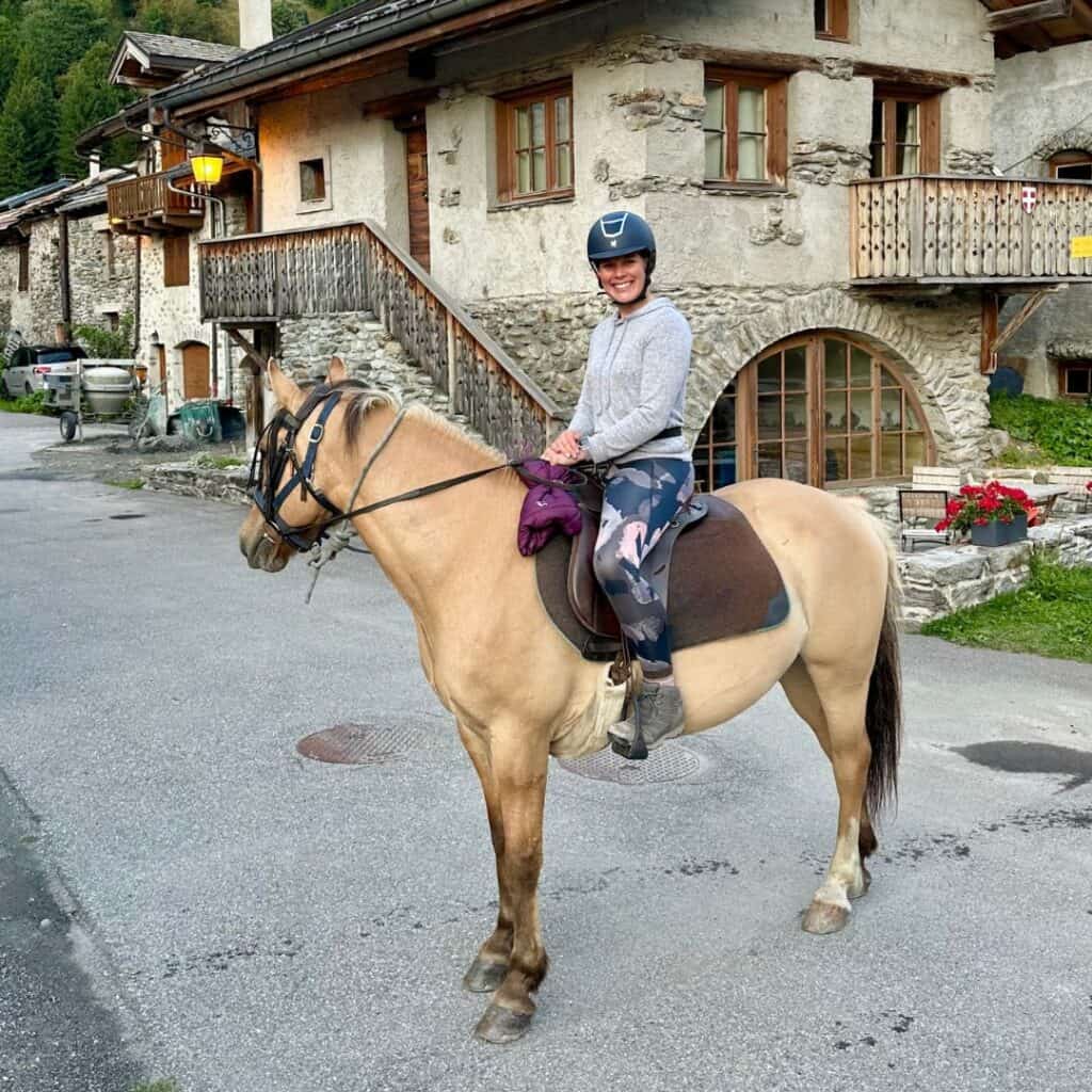 A woman sits confidently on a light brown horse in a charming stone village near Chalet des Vignobles. Old alpine architecture with wood balconies frames the peaceful setting.