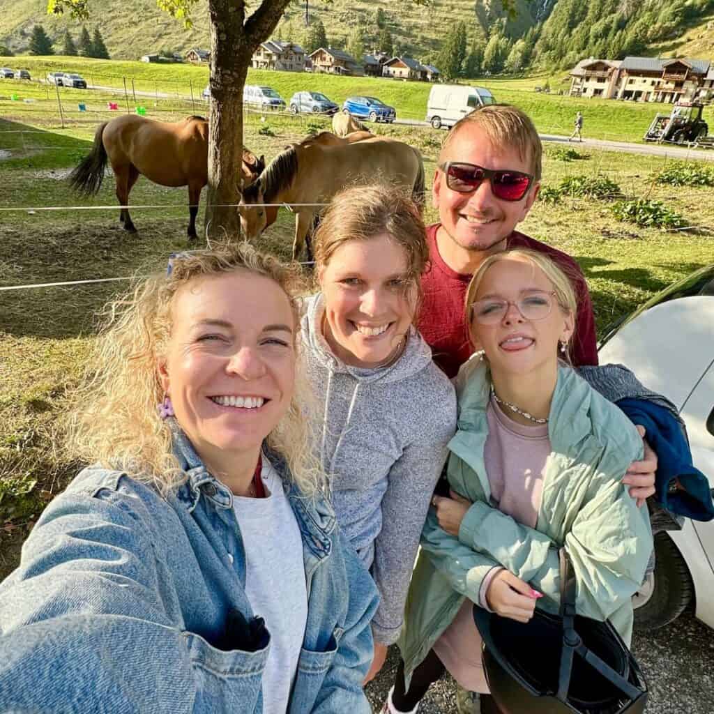 Four smiling friends pose for a group selfie in front of grazing horses in a fenced paddock. The sunny weather and alpine backdrop add to the joyful atmosphere of their La Plagne horse riding adventure.