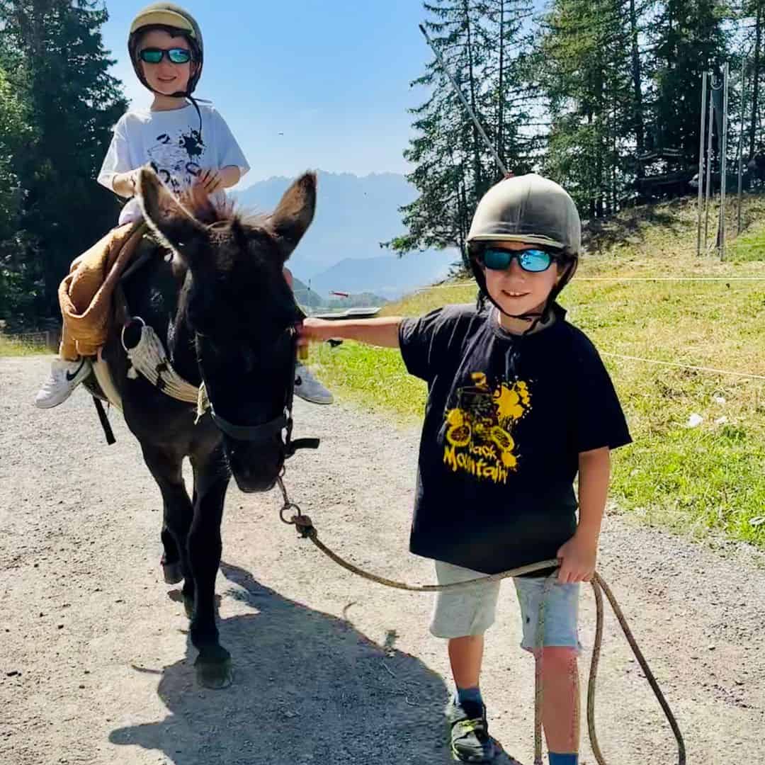 Two children in helmets and sunglasses enjoy pony trekking in La Plagne, with one sitting on a black donkey and the other holding the lead rope. Mountain views peek through the trees under a bright summer sky.