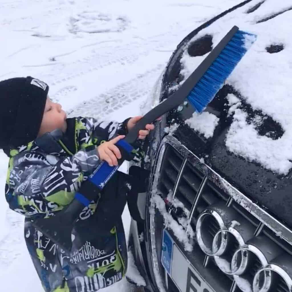 A bundled-up toddler uses a snow brush to clear snow off the hood of a parked Audi, revealing part of a smiley face drawn in the snow. The snowy ground and winter gear highlight typical ski holiday conditions, subtly nodding to the importance of car prep and travel readiness with upcoming EES border changes.