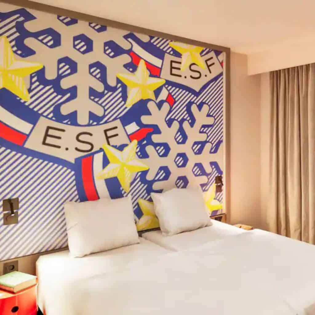 Modern hotel room with two twin beds pushed together under a large, colorful pop-art mural featuring snowflakes, yellow stars, and the letters “E.S.F.” on a bold striped background. The playful design reflects the vibrant atmosphere of PopAlp Hotel in Plagne Centre.