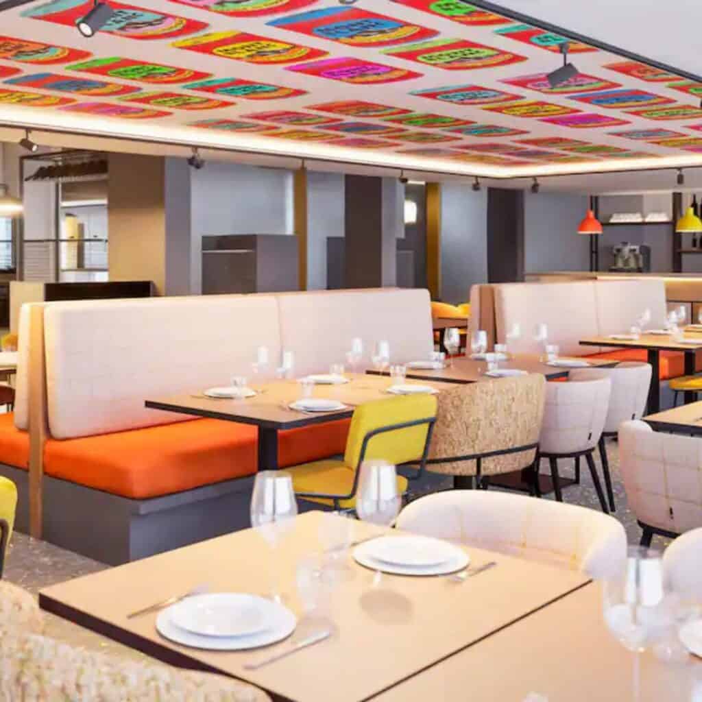 Bright and cheerful dining area with orange and yellow accents, booth seating, and neatly set tables. The PopAlp Hotel restaurant in Plagne Centre features bold ceiling art with repeating colorful graphic patterns.