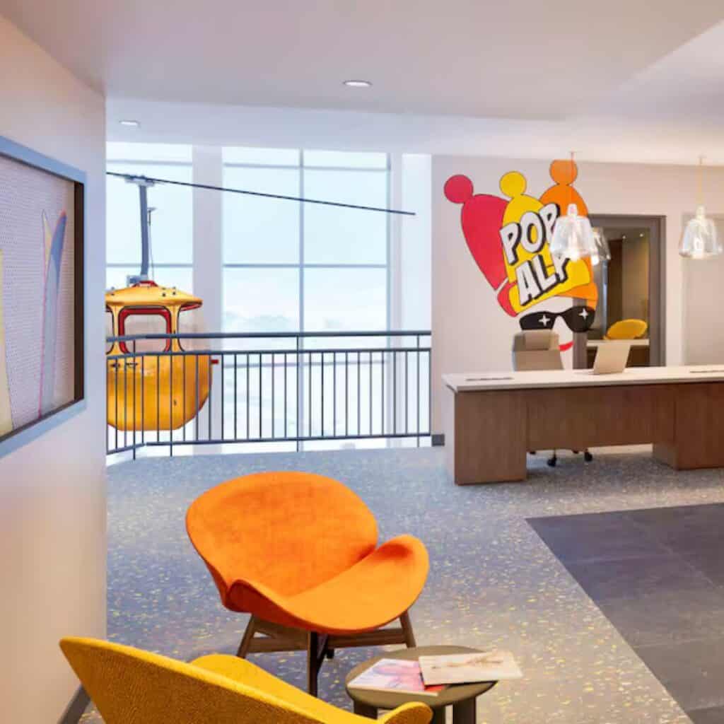 Lobby area with bright orange chairs, floor-to-ceiling windows, and a whimsical wall graphic that says “Pop Alp” in bold comic-style lettering. A yellow ski gondola is visible outside, emphasizing the mountain theme of the PopAlp Hotel in Plagne Centre.