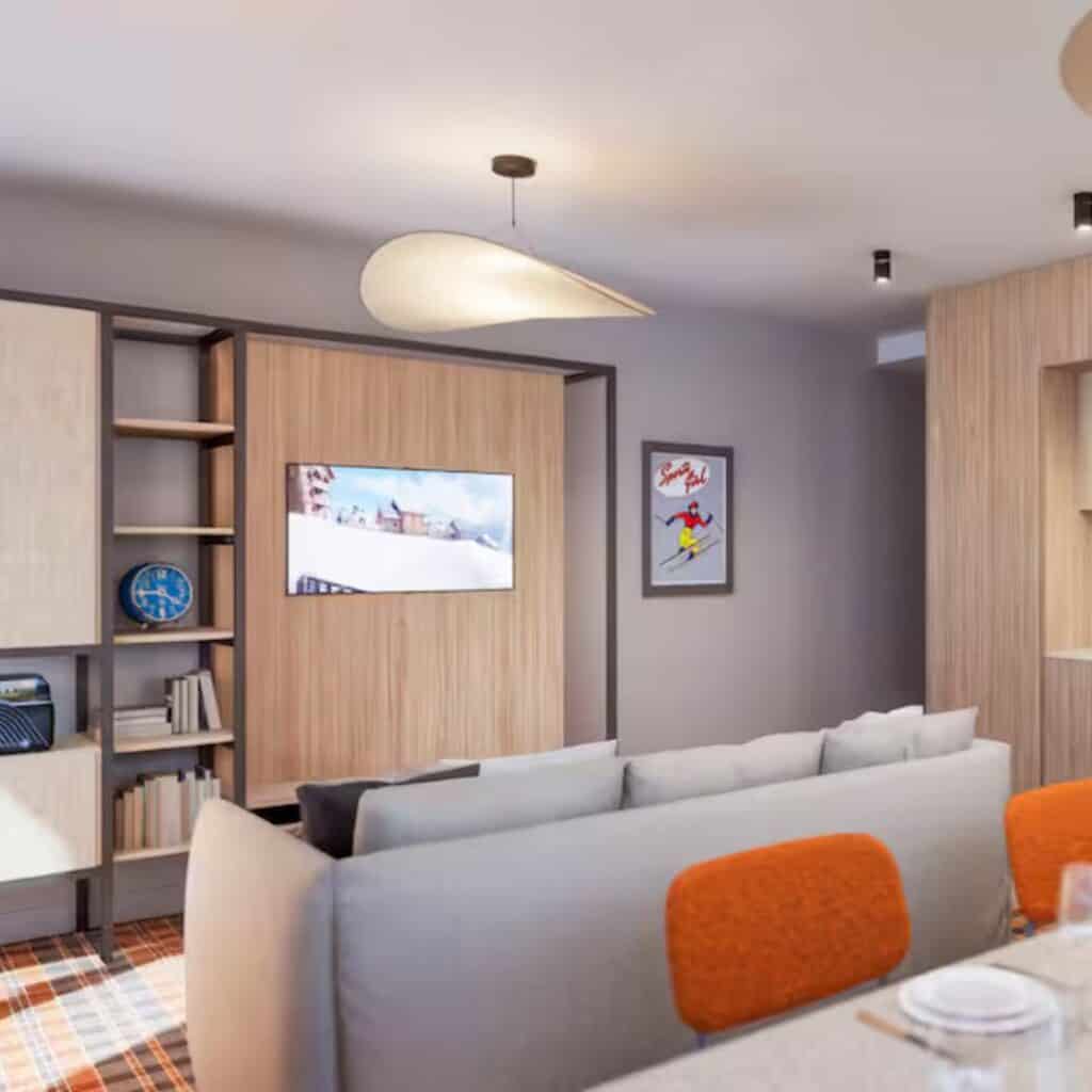 Contemporary suite lounge area with a cozy grey couch, orange-backed chairs, and wood paneling surrounding a flat-screen TV and ski-themed wall art. The modern alpine decor ties into the stylish comfort offered by PopAlp Hotel in Plagne Centre.