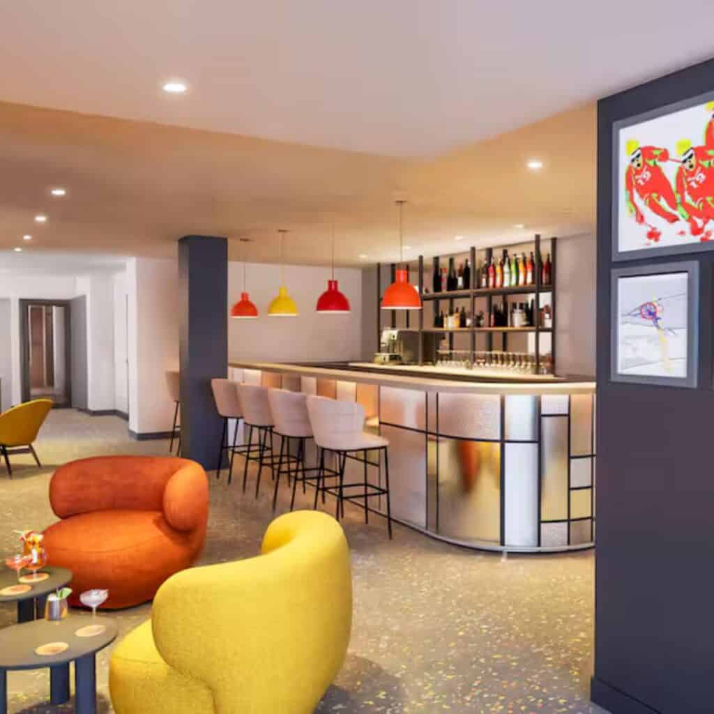 Colorful hotel bar with high stools, a sleek counter, and pendant lights in red, yellow, and orange. The lounge at PopAlp Hotel in Plagne Centre is lively and welcoming, with quirky art and cozy seating areas.