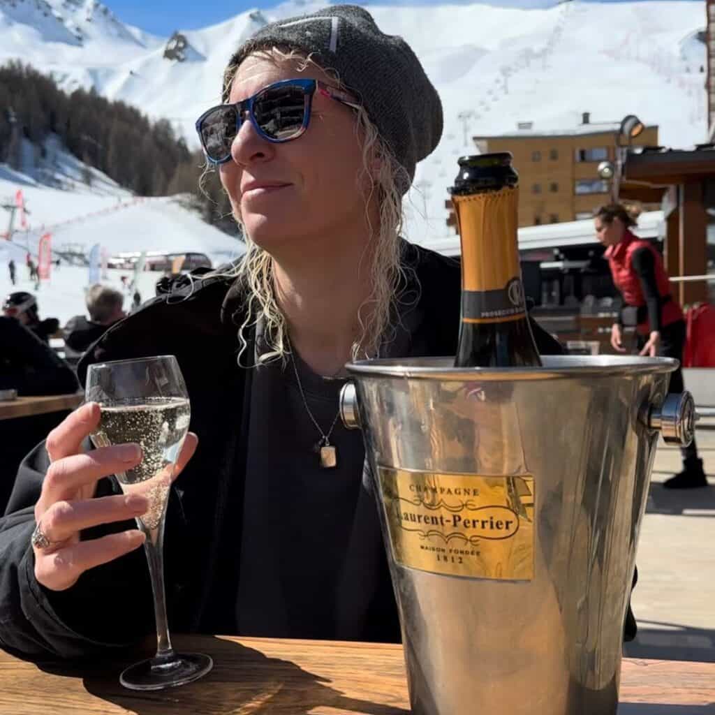 Woman in sunglasses and a beanie enjoys a glass of sparkling wine on a sunny terrace in La Plagne, with a bottle of Laurent-Perrier chilling in an ice bucket and snowy ski slopes in the background.