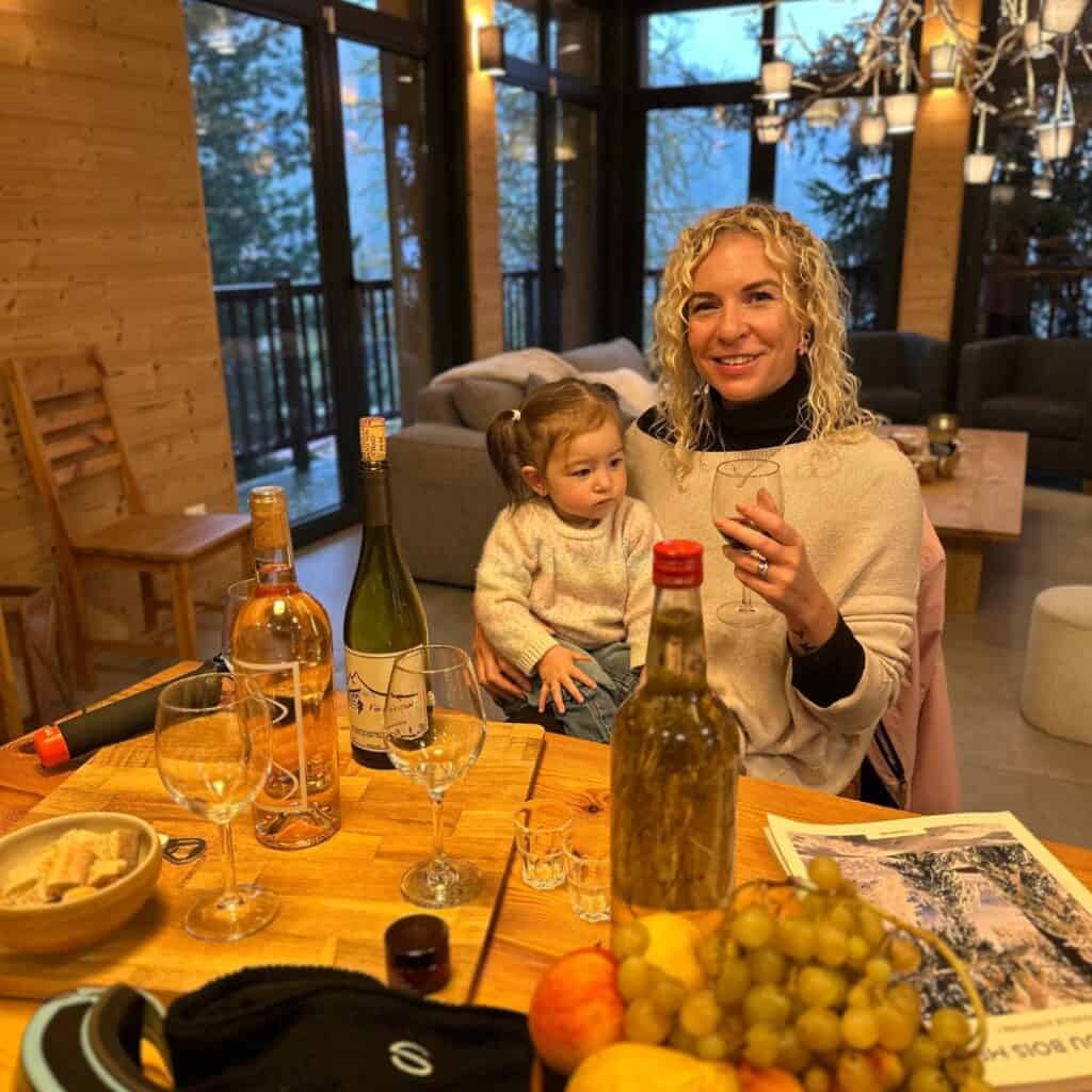 Smiling woman holding a glass of red wine while sitting at a chalet table with a toddler on her lap, surrounded by bottles of local wine, fruit, and a cozy mountain view through large windows.