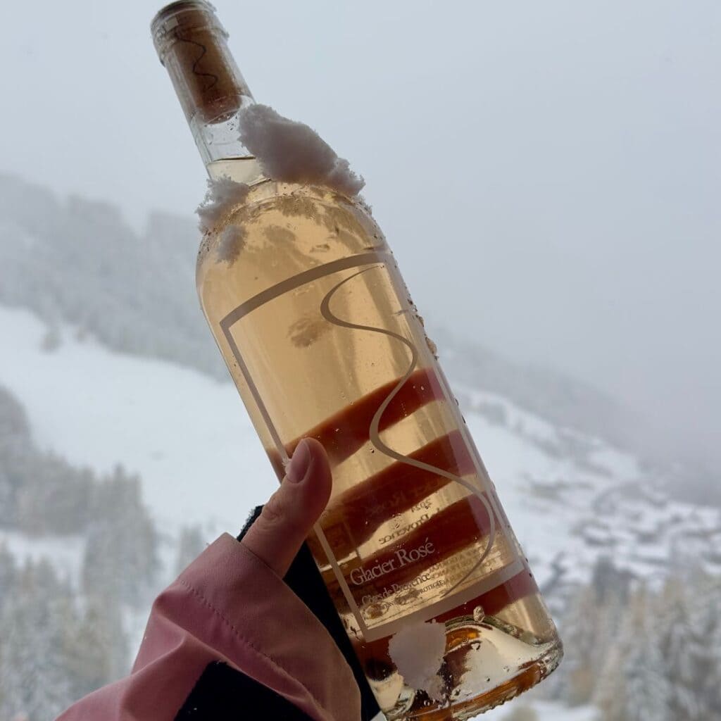 Hand holding a bottle of Glacier Rosé wine dusted with snow, with a misty, snow-covered mountain landscape in the background.