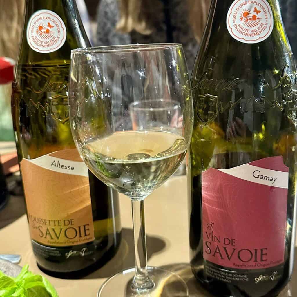 Two bottles of Savoie wine—Roussette de Savoie Altesse and Vin de Savoie Gamay—flank a glass of white wine on a table during a tasting.