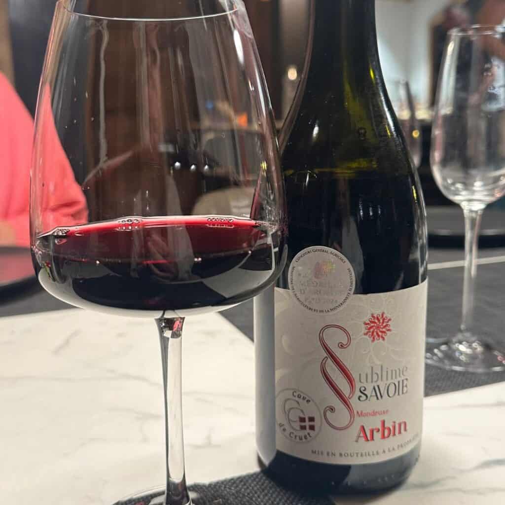 A deep red glass of Mondeuse wine sits beside a bottle labeled Sublime Savoie Arbin, on a marble table at a cozy dinner.