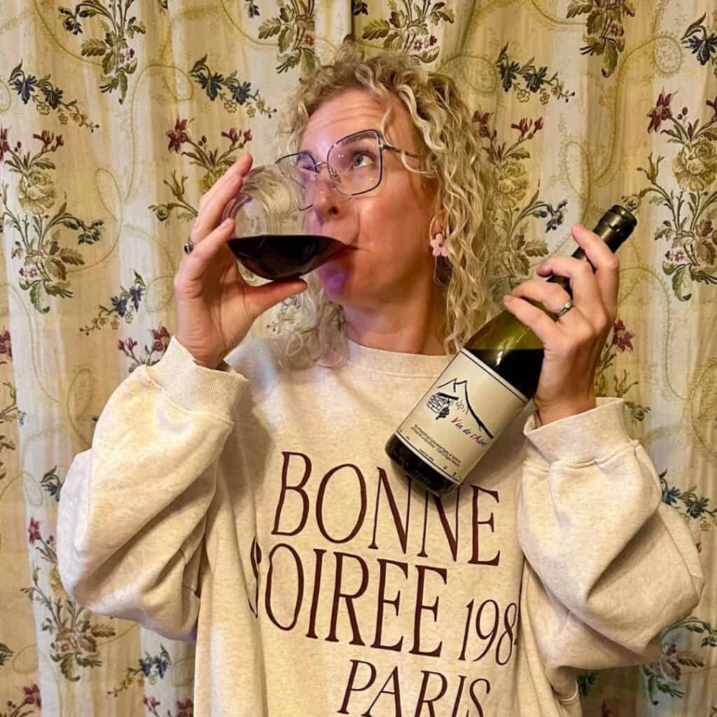 A woman in a “Bonne Soirée Paris” sweatshirt drinks red wine straight from a glass while holding a bottle of Vin de l’Adret.